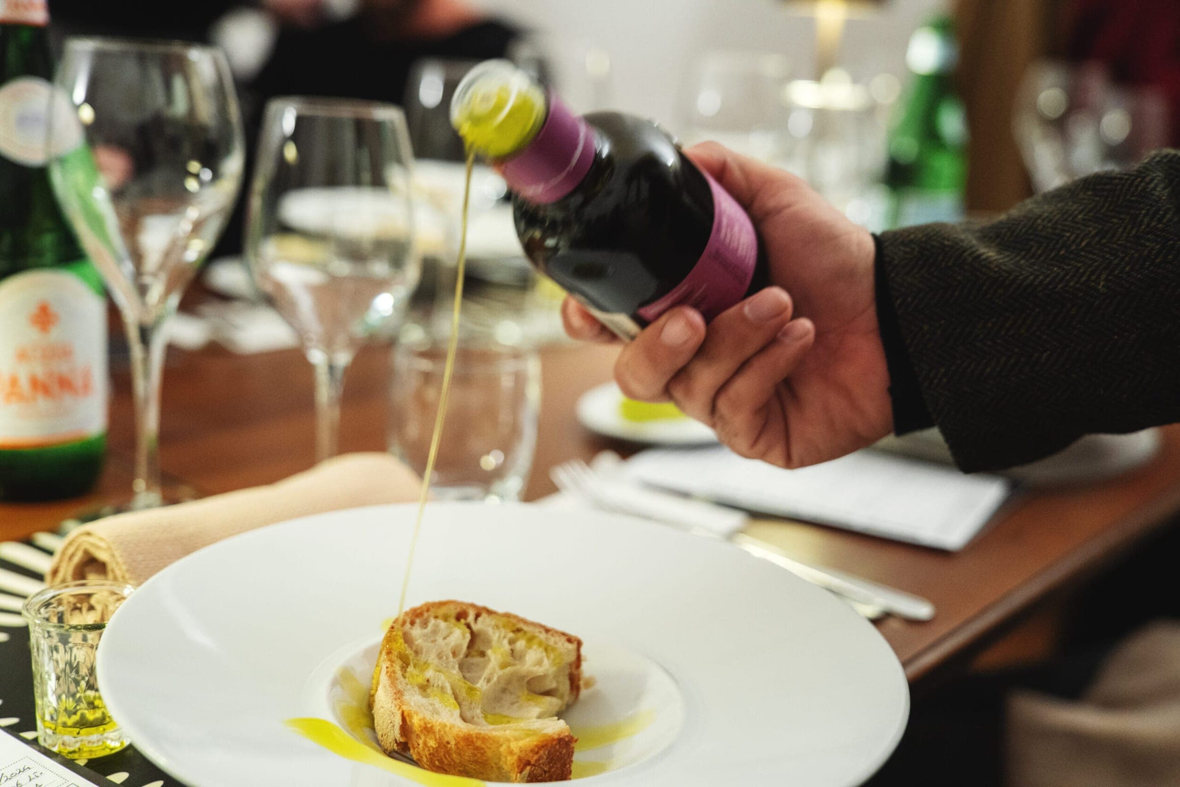 Olio extravergine versato su una fetta di pane, con bicchieri e un tavolo apparecchiato sullo sfondo