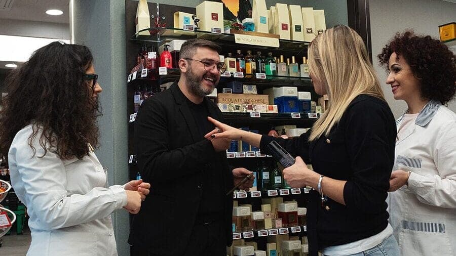 In farmacia, un uomo in giacca nera conversa con una donna che tiene in mano un prodotto, tra due farmaciste in camice bianco
