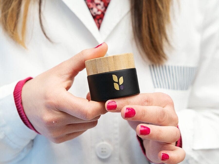 Un barattolo nero con il tappo in bambù e il logo a foglie, tenuto tra le mani da una farmacista in camice
