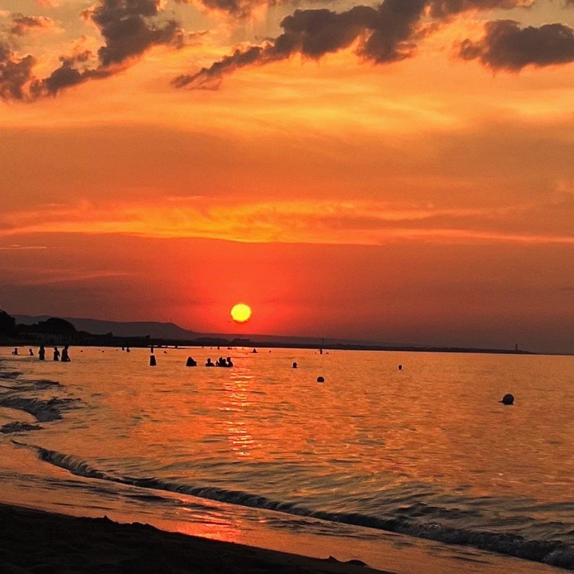 Il sole arancione tramonta sull’orizzonte del mare; in acqua, vicino alla riva, le sagome di alcuni bagnanti