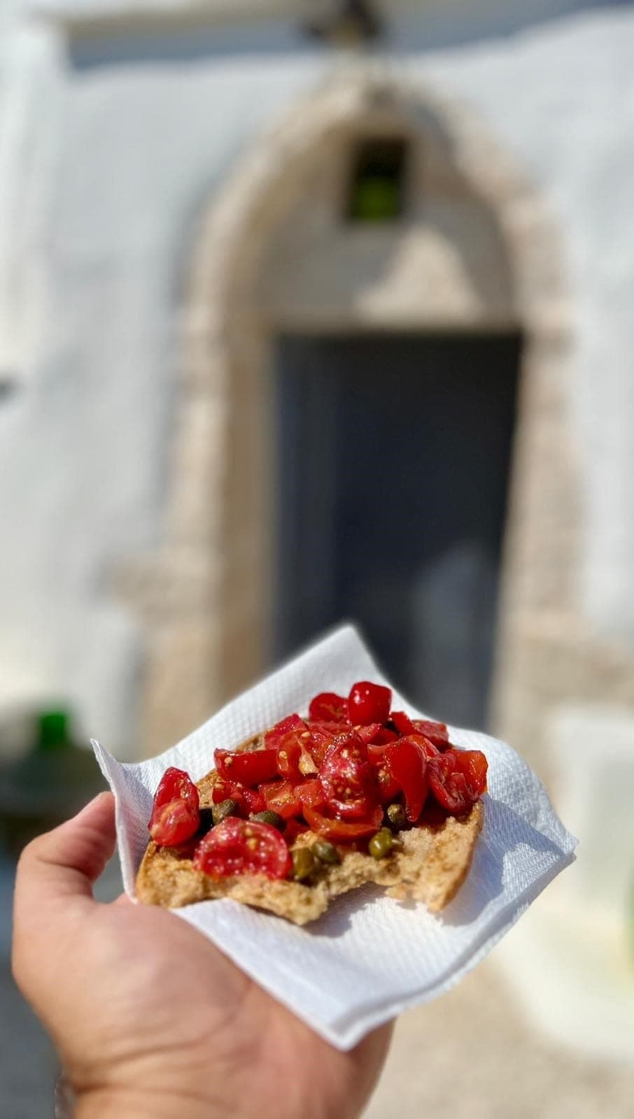 Una mano tiene una fetta di pane tostato con pomodorini e capperi, davanti alla porta del trullo