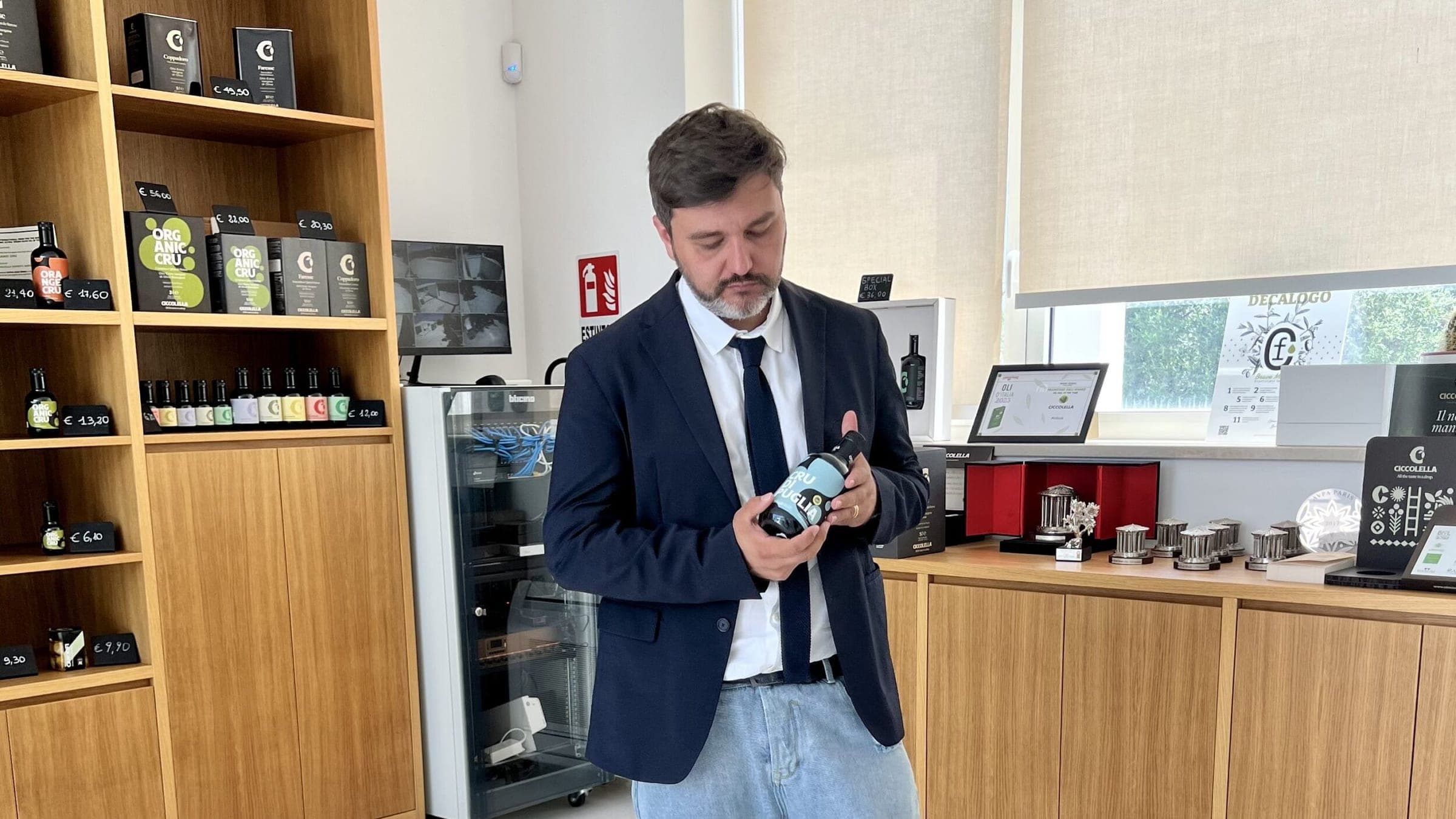 Un uomo in blazer blu esamina una bottiglia di Cru di Puglia nello spazio vendita del frantoio, tra scaffali di legno