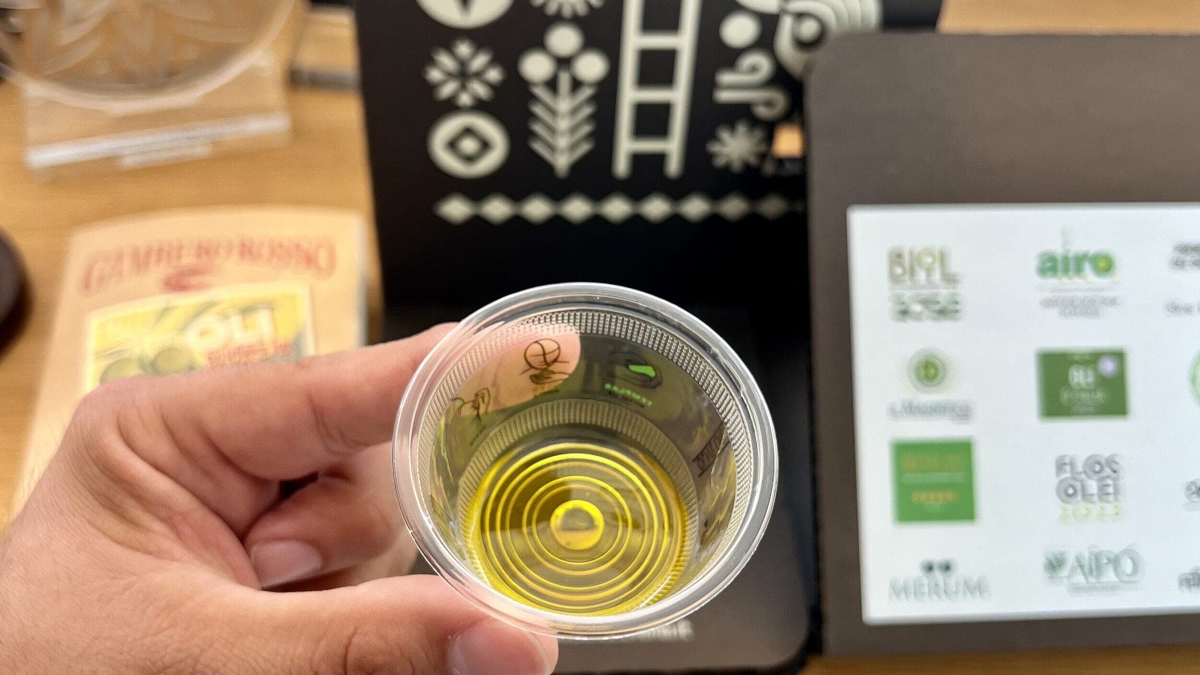 Una mano regge dall’alto un bicchierino con olio giallo-verde, davanti a una confezione scura e a un pannello di premi