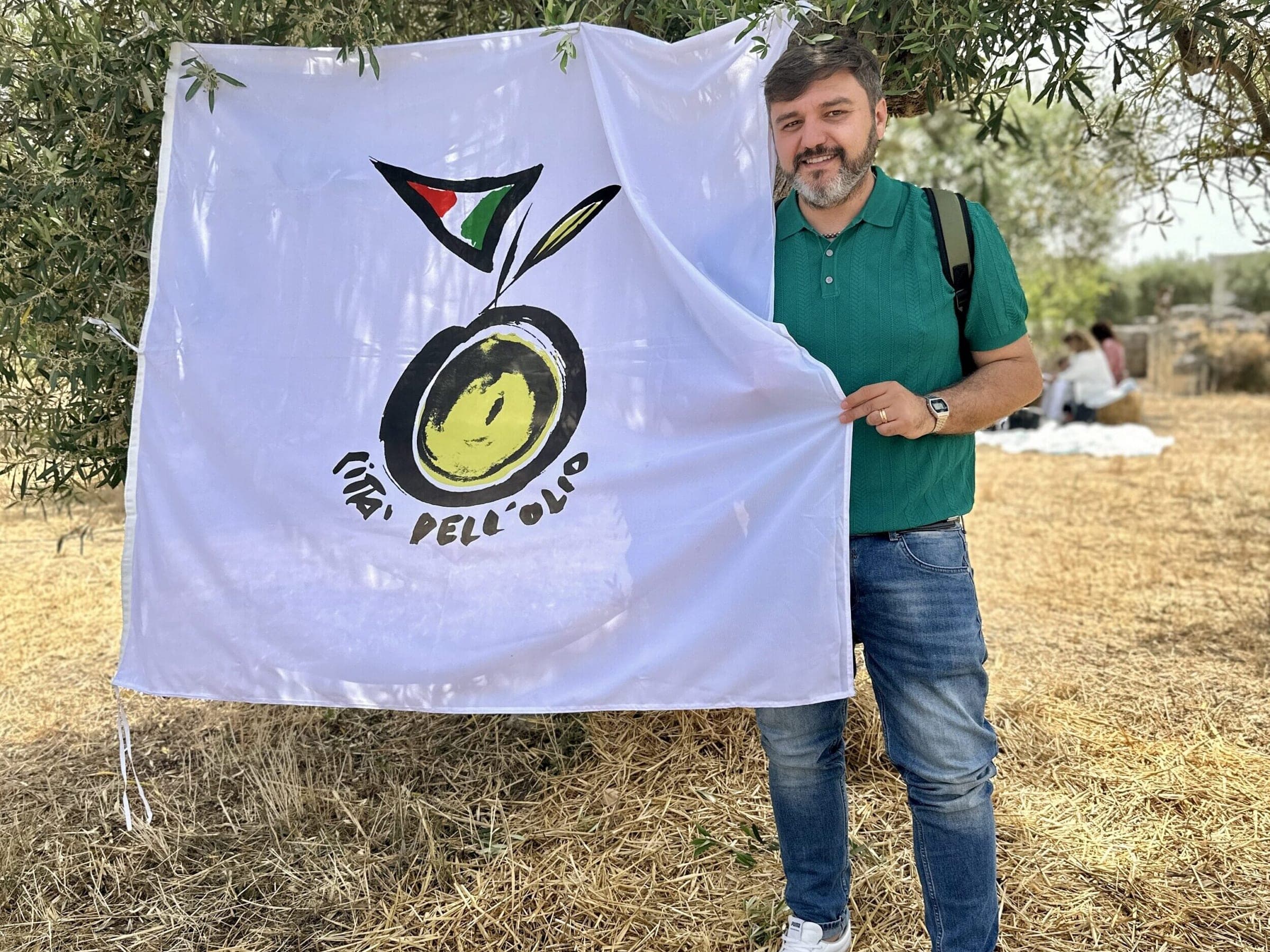 Un uomo in polo verde sorride accanto alla bandiera di Città dell’Olio appesa a un ulivo; sullo sfondo, un picnic
