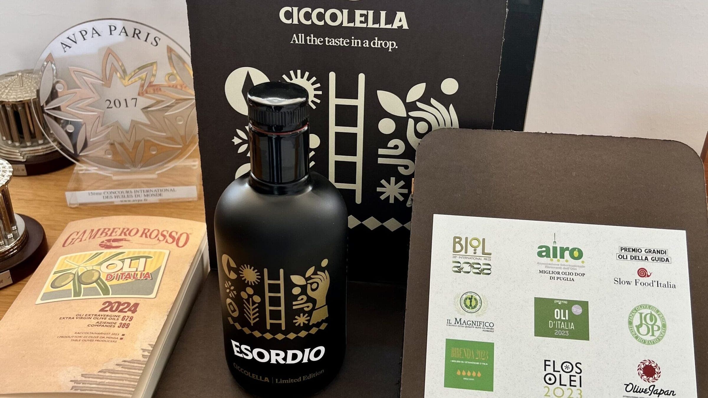 La bottiglia nera Esordio davanti a una busta scura, con la guida Oli d’Italia 2024 e un trofeo AVPA Paris accanto