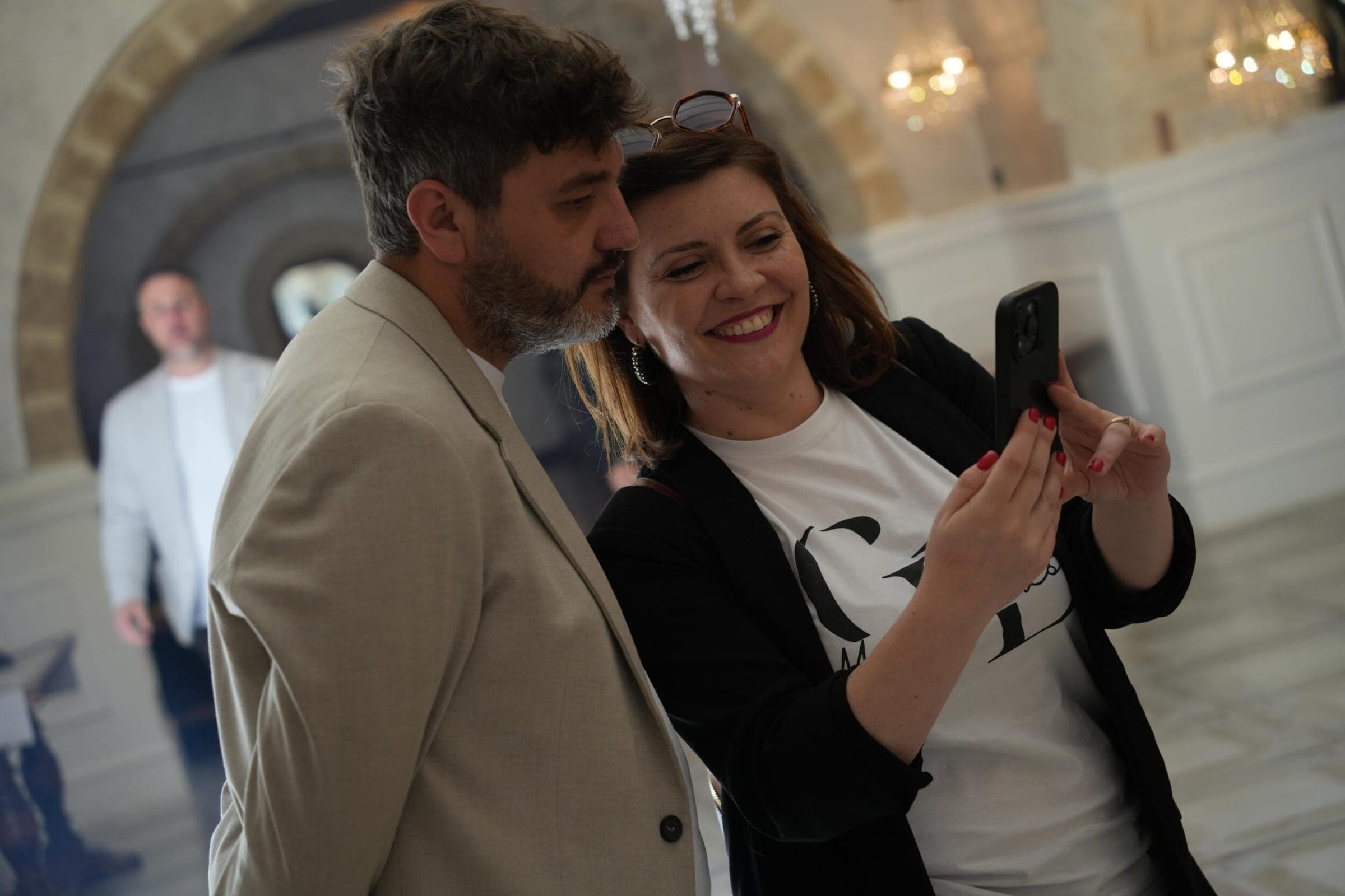 Matteo Contino e una donna sorridente guardano insieme un telefono, sotto un arco di pietra