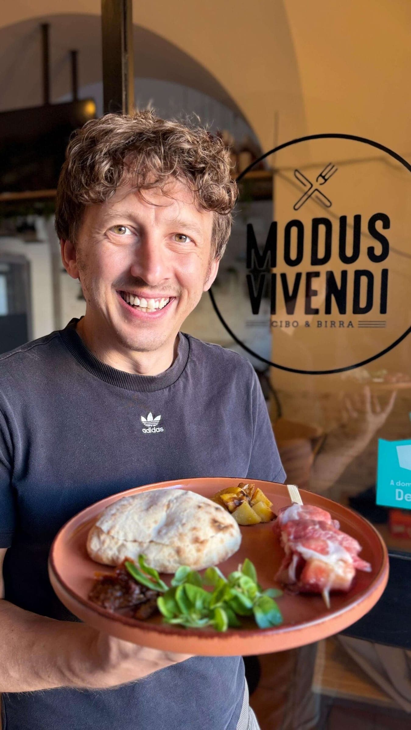 Modus Vivendi Cibo e Birra: un uomo sorridente tiene un piatto con pane e salumi